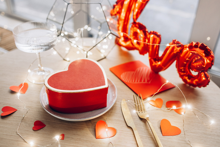Romantic table setting with a heart-shaped gift box on a plate, surrounded by golden cutlery, red heart decorations, and warm string lights, with a "love" balloon in the background, creating a cozy and festive atmosphere