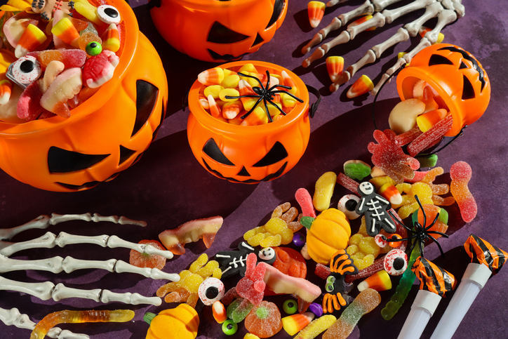 Image of four plastic, orange Jack O'Lantern pumpkin sweet collection buckets, trick or treat containers full of gummy sweets, surrounded by spilled sweet and sour jellied candy, candy corn, skeletal hands, spiders, purple background, elevated view