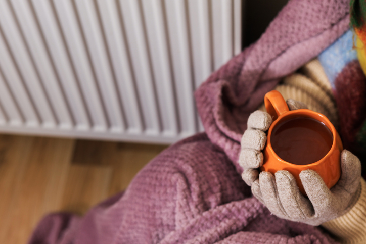 A close-up of a girl holding a cup of tea. She sits near a radiator. She's wearing a scarf, and a sweater.